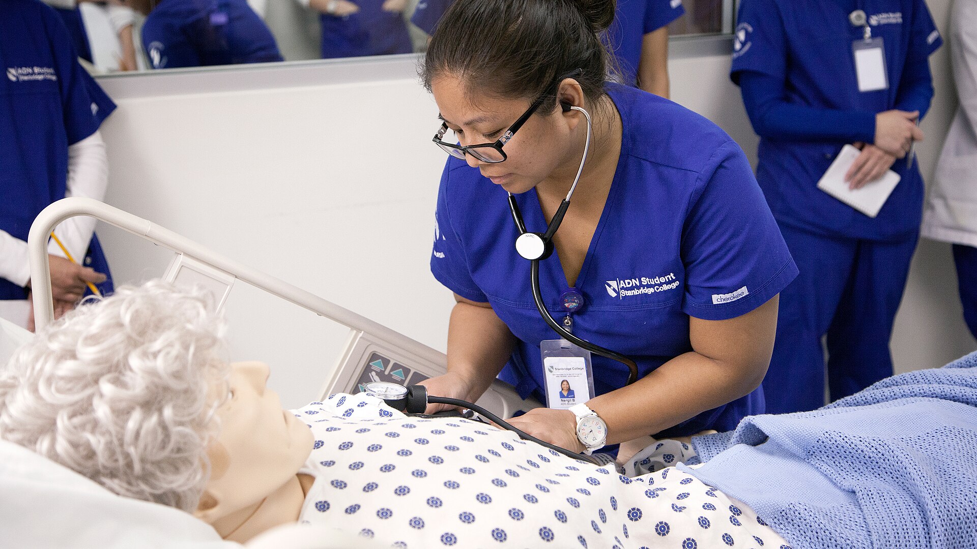 A nursing student in blue scrubs takes a manikin's blood pressure in a simulation lab while classmates watch, the training that comes before paying the NCLEX exam cost.