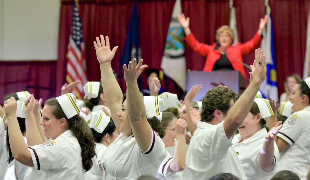 Nursing graduates in white uniforms and caps raise their hands and cheer at a pinning ceremony while a speaker in red raises her arms at the podium.