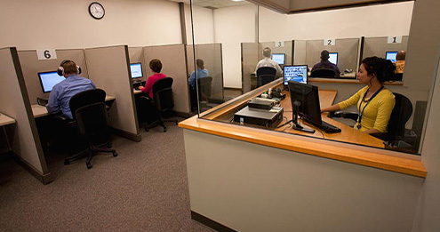 A Pearson Professional Center testing room with numbered computer booths and a proctor seated at a glass-walled station.