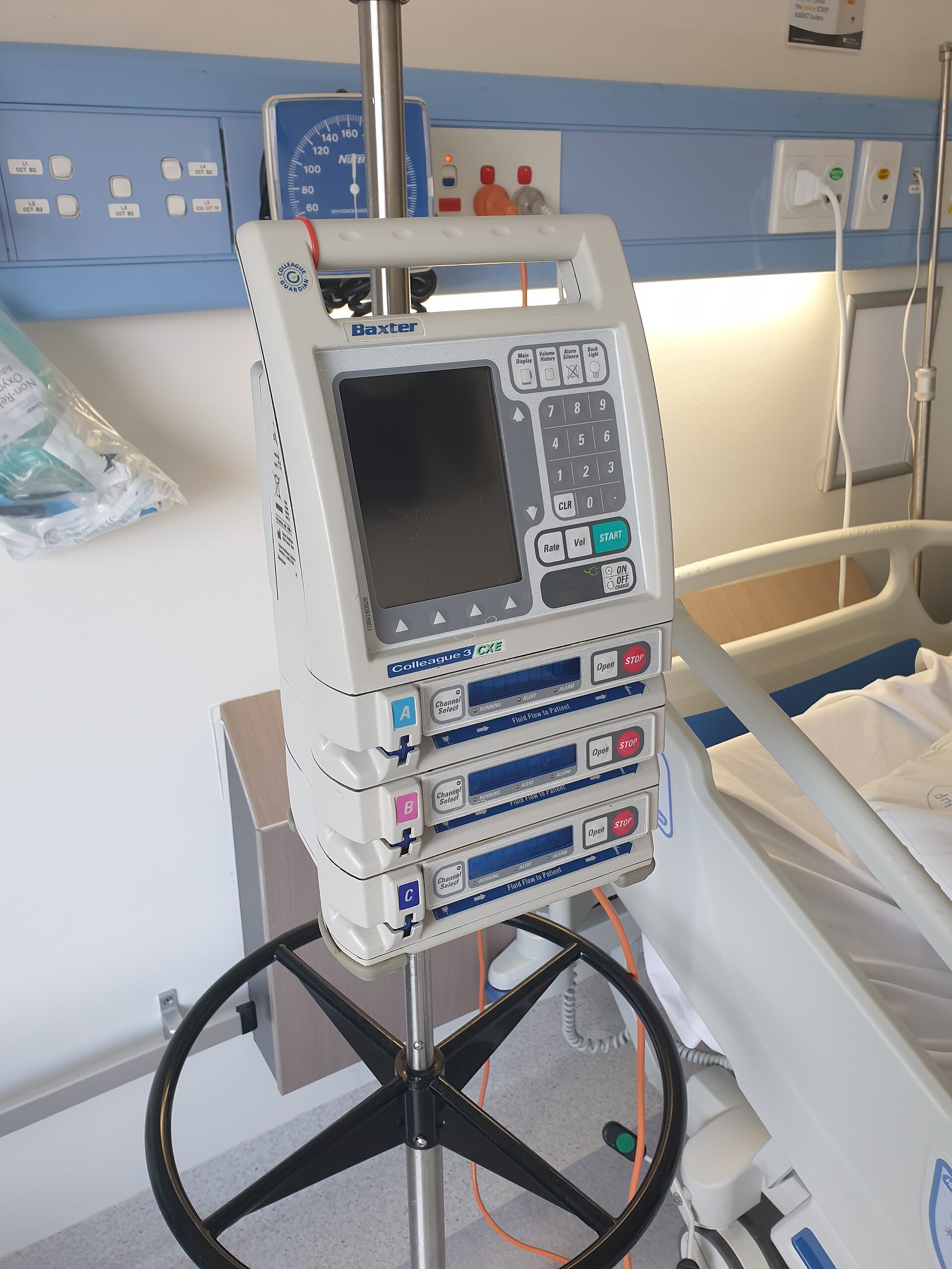 A three-channel IV infusion pump mounted on a pole next to a hospital bed, with a wall-mounted blood pressure gauge behind it.
