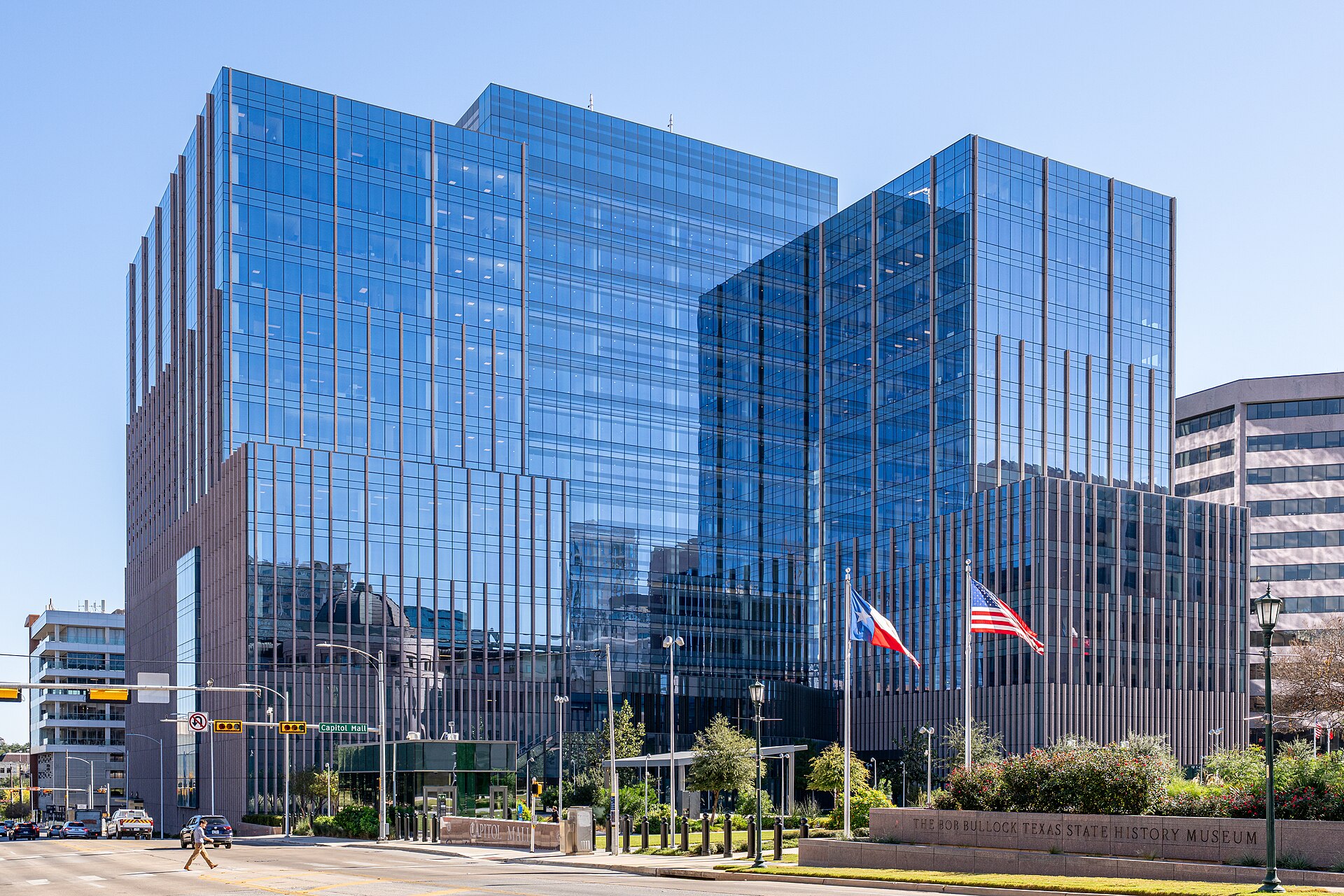 The glass-walled George H. W. Bush State Office Building on Congress Avenue in Austin, Texas, with Texas and U.S. flags in front.